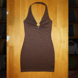 Brun zebramönstrad halterneckklänning - Snygg kort brun klänning med zebramönster och halterneck. Klänningen har en ringdetalj framtill vid bysten och är figurnära med stretchigt material. Perfekt för dig som gillar trendiga mönster och vill sticka ut.