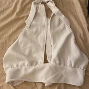 Vit halterneck linnetopp med knyt - Snygg vit halterneck linnetopp med bred resår nedtill och knyt i nacken. Toppen har en djup v-ringning framtill och är ärmlös. Enkel och stilren design som passar till mycket.