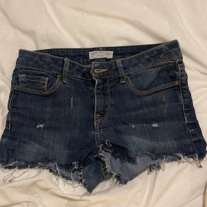 Mörkblå jeansshorts från Denim Brunex - Snygga mörkblå jeansshorts från Denim Brunex med råa, fransiga kanter och slitna detaljer. Klassisk femficksmodell med orange kontrastsömmar och låg midja. Perfekta för sommaren och ger en cool, avslappnad vibe. Upskattar till en strök XS-S