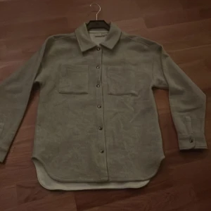 Grå overshirt från Name It - Stilren beige overshirt från Name It med klassisk krage, två bröstfickor och knappar framtill. Skjortan har långa ärmar med knapp vid ärmslut och är tillverkad i ett mjukt material som känns skönt mot huden. Perfekt för lager-på-lager-stil.