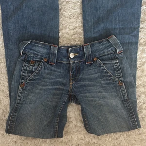 True Religion jeans från 2000-talet - Säljer ett par blå jeans från True Religion med ikoniska broderade bakfickor och patch med logga. Jeansen har slitna detaljer och råa benslut för en edgy look. Klassisk femficksmodell med orange kontrastsömmar och knappgylf.