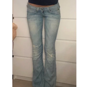 True Religion ljusblå bootcut jeans - Säljer ett par ikoniska ljusblå jeans från True Religion med bootcut passform och låg midja. Jeansen har slitna detaljer, klassiska gula sömmar och snygga bakfickor med broderad logga. Perfekta för dig som gillar vintagevibbar och streetstyle.