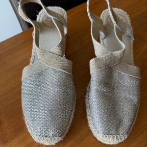 Toni Pons sandaler med platå, strl 39 - Snygga sandaler från Toni Pons i storlek 39 med glittrig silverfärgad ovandel och breda band i beige textil. Skorna har en rejäl platåsula i flätad jute och öppen häl. Perfekta för sommaren när du vill sticka ut lite extra.