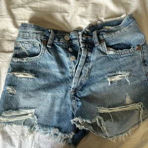 Blå slitna jeansshorts från Zara - Säljer ett par blå jeansshorts från Zara i midwaist-modell. Shortsen har flera slitningar och fransiga benslut för en riktigt cool och avslappnad vibe. Klassisk femficksdesign och knäppning med knappar framtill. Perfekta för sommardagar när du vill ha en edgy look.