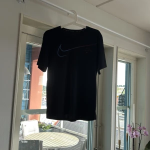 Svart Nike t-shirt med logga - Svart t-shirt från Nike med klassisk swoosh-logga i blått och rött framtill. Enkel och sportig design med rund hals och korta ärmar. Perfekt för dig som gillar en clean look och sportiga vibes.