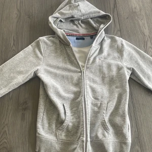 Hoodie - Snygg ljusgrå hoodie från Gant med dragkedja och huva. Tröjan har två stora fickor framtill och klassisk Gant-logga på bröstet. Perfekt för chill dagar och enkel att matcha med jeans eller joggers.