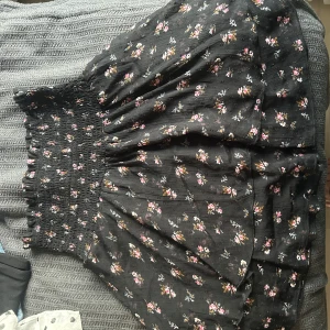 Svart blommig kortkjol med smock - Supersöt svart kortkjol med små blommor i rosa, vitt och gult. Kjolen har bred smock i midjan som ger en skön och flexibel passform. Tyget är lätt och stretchigt.