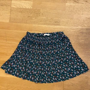 Blommig kortkjol - Kortkjol med småblommigt mönster i lila, rött och vitt på mörkgrön botten. Kjolen är plisserad och har resår i midjan. Bekväm och i väldigt bra skick! 