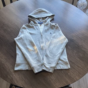 Grå hoodie från Polo Ralph Lauren - Snygg grå hoodie från Polo Ralph Lauren med dragkedja och klassisk blå broderad logga på bröstet. Tröjan har huva, långa ärmar och känguruficka framtill. Mjuk insida i bomullsblandning, perfekt för chill dagar.
