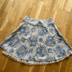 Blåmönstrad kjol från Vero Moda - En kjol från Vero Moda med beige botten och blått blommönster. Kjolen har quiltad midja, små paljettdetaljer och en volang av tyll längst ner. Några paljetter kan saknas men det är inget man märker. Hör av dig vid frågor! 😊