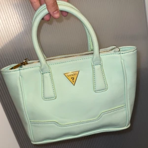 Guess vintage mintgrön väska - Superfin mintgrön handväska från Guess med klassisk form och guldfärgade detaljer. Väskan har två rejäla handtag, dragkedja upptill och ett rymligt innerfack med extra ficka. Tillverkad i syntetmaterial med stilren look och diskret logga framtill. Använd endast ett fåtal gånger så den är i bra skick. Priset kan diskuteras.