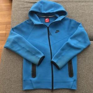 Blå Nike hoodiejacka med dragkedja XS - Nike hoodiejacka i klarblå färg med svarta detaljer och hel dragkedja. Jackan har två fickor med dragkedja, huva och ribbade muddar. Perfekt för dig som gillar sportig och clean stil. Snygg Nike-logga på bröstet.