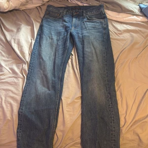 Blå jeans från Nudie Jeans, W28 L32 - Säljer ett par klassiska blå jeans från Nudie Jeans med raka ben och snygga kontrastsömmar. Jeansen har fem fickor, normal passform och är tillverkade i ekologisk bomull. Bakfickorna har Nudie Jeans ikoniska broderi. Köpta för 1699 bra skick kom med frågor