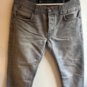 Grå jeans från Crosshatch W32 - Snygga grå jeans från Crosshatch med klassisk femficksdesign och röda sömdetaljer. Jeansen är tillverkade i en mix av bomull och polyester för en skön känsla och bra passform. Perfekta för dig som gillar en stilren look.