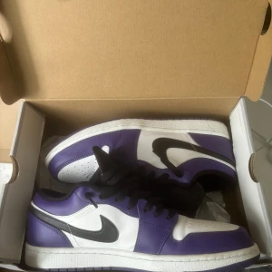 Nike Air Jordan 1 Low Court Purple - Säljer ett par Nike Air Jordan 1 Low i färgen Court Purple. Skorna har en snygg kombo av vitt, svart och lila läder, med klassisk svart swoosh och svarta skosnören. Låg modell med perforerad tå och ikonisk Jordan-logga på plösen.