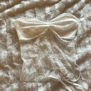 Vit spets tubtopp med snörning - Supersöt vit tubtopp i spets med transparent blommönster och snygg snörning baktill. Toppen har bandeau-modell och är perfekt för att skapa en romantisk och trendig look. Tunn och luftig, passar perfekt till sommarens outfits.