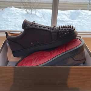 Christian Louboutin grå sneakers med nitar - Grå sneakers från Christian Louboutin i mocka med ikoniska röda sulor och grå nitar på tån. Klassisk låg modell med snörning och rund tå. Snygg och edgy design som sticker ut, perfekt för dig som vill ha något extra.