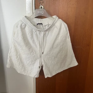 Vita Louis Vuitton shorts med monogram - Snygga vita shorts från Louis Vuitton med ikoniskt monogrammönster i tyget. De har elastisk midja med snörning och är tillverkade i ett mjukt bomullsmaterial / Handduksmaterial. Perfekta för en clean och lyxig look, passar till allt från chill till streetstyle. Inget kvitto och därav priset