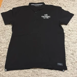 Svart Peak Performance pikétröja XL - Svart pikétröja från Peak Performance i storlek XL. Klassisk krage med knappar och vit logga på bröstet. Tillverkad i mjukt material, perfekt för en clean och sportig look. Diskreta ränder på insidan av kragen och liten etikett nertill.