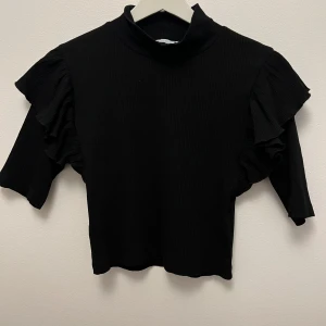 Svart volangtop från Zara - Svart ribbad topp från Zara med höghalsad krage och snygga volangdetaljer på axlarna. Toppen har halvlånga ärmar och en loose passform som ger en cool och trendig look. Blusen är i storlek L men skulle säga att den passar en S.