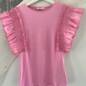 Rosa volangblus från Amisu - Supersöt rosa blus från Amisu med korta ärmar och breda volanger i glansigt tyg längs axlarna. Toppen har en rund halsringning och smockdetaljer på ärmarna som ger en unik och trendig look. Perfekt för dig som vill sticka ut med färg och volym.