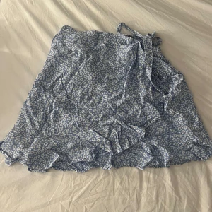 Blommig omlottkjol från Indiska - Supersöt blå omlottkjol från Indiska i storlek S med vitt blommönster och volangkant. Kjolen har knytband i midjan och är perfekt för en söt sommarlook. Köpt för ca 500kr