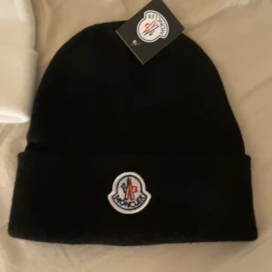 Svart Moncler mössa med logga - Svart mössa från Moncler med klassisk uppvikt kant och broderad Moncler-logga framtill. Enkel och stilren design som passar perfekt till kalla dagar. Mössan är tillverkad i mjukt material för extra komfort. FINNS ÄVEN I VIT