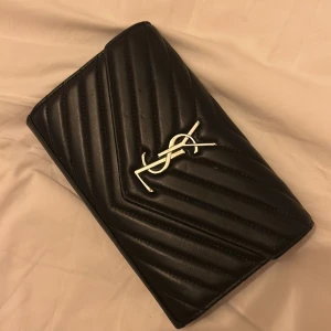 Svart quiltad YSL axelväska i skinn - Säljer en svart quiltad axelväska från YSL med silverfärgad kedja och ikonisk YSL-logga framtill. Väskan är tillverkad i äkta läder och har ett elegant, stilrent mönster med snedställda sömmar. Perfekt storlek för det viktigaste och ger en lyxig vibe till din outfit. Knappt använd.