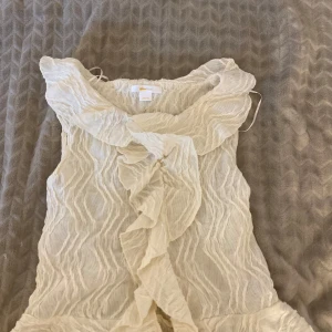 Vit volangblus från Gina Tricot - Supersöt vit blus från Gina Tricot med volangdetaljer längs halsringningen och framtill. Tunt, vågigt tyg i luftig bomull som ger en romantisk vibe. Perfekt för dig som gillar feminina och trendiga toppar med extra detaljer.