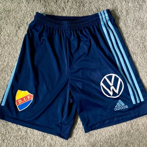 Blå DIF Adidas shorts 176 eller S - Snygga marinblå shorts från Adidas med Djurgårdens IF-logga och Volkswagen-tryck. Klassiska ljusblå ränder längs sidorna och elastisk midja. Perfekta för fotbollsträning eller chill. Tillverkade i polyester för skön känsla och bra rörelsefrihet.