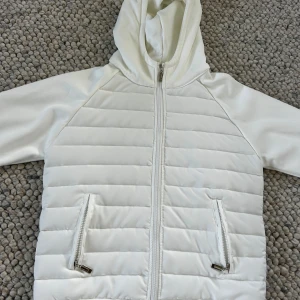 Vit cardigan med huva oanvänd  - Snygg vit Cardigan med quiltad framsida och huva. Jackan har dragkedja framtill och två fickor med dragkedja. Perfekt för kyliga dagar när du vill hålla stilen enkel och fräsch.