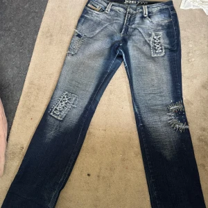 Blå jeans y2k  - Säljer ett par blå jeans från Jeans PYP med slitna detaljer och snörning på både fram- och bakfickor. Jeansen har en tvättad look, raka ben och coola nitar runt snörningen. Perfekta för dig som gillar en edgy stil.