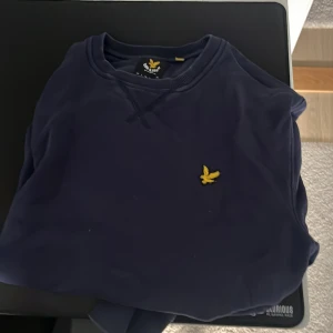 Marinblå sweatshirt från Lyle & Scott - Säljer en marinblå sweatshirt från Lyle & Scott med klassisk gul logga broderad på bröstet. Tröjan har rund halsringning, ribbade muddar och är tillverkad i mjuk bomull. Perfekt för dig som gillar stilren och enkel design.