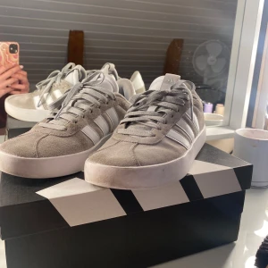 Gråa Adidas sneakers samba i mocka - Snygga gråa Adidas sneakers med klassiska vita ränder på sidorna. Ovandelen är i mjuk mocka och skorna har vita detaljer och snörning. Perfekta för en clean och stilren look, passar till det mesta i garderoben. OBS: hål på vänstra sko. Syns inte om man inte kollar nära (se på bild 3) ❤️