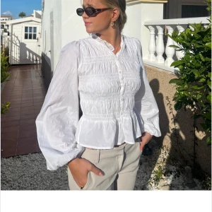 Vit blus med smock och volang - Säljer en vit blus med smockdetaljer över bröstet och volang nertill. Blusen har långa puffärmar och en liten ståkrage. Perfekt för dig som gillar romantisk och stilren look. Storlek 36