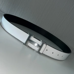Vändbart bälte med H-spänne - Stilrent vändbart bälte i skinn med en sida i vitt och en sida i svart. Bältet har ett stort H-format spänne i silverfärg och klassisk design. Perfekt accessoar för att lyfta din outfit.