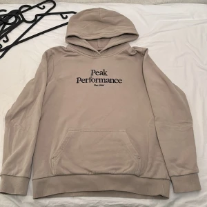 Beige hoodie från Peak Performance - Säljer en beige hoodie från Peak Performance med broderad logga på bröstet. Tröjan har huva, magficka och ribbade muddar vid ärmslut och nederkant. Perfekt för en avslappnad stil.