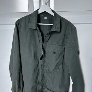 Grön overshirt från C.P. Company - Snygg grön overshirt från C.P. Company med dragkedja och knappstängd bröstficka. Klassisk krage och ikonisk linsdetalj på ärmen. Väldigt tunt o luftigt material, perfekt lager-på-lager-plagg med modern känsla. Strl M men har du S så funkar de på dig också. Hör av er vid intresse/ frågor!