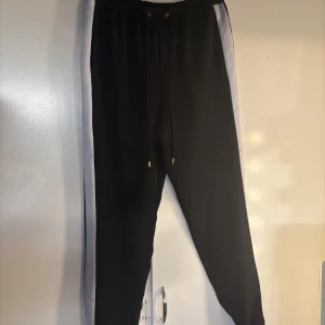 Svarta byxor från Michael Kors med vita sidor - Svarta byxor från Michael Kors med vita paneler längs sidorna och guldfärgade detaljer. Byxorna har resår i midjan med snörning och fickor fram. Använda en gång. Köpta på Zalando 