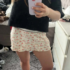 Vit kjol med körsbärsmönster från Zara - Supersöt vit kjol från Zara med rött körsbärsmönster och volangdetaljer. Kjolen är kort och har en lätt, flowy känsla som passar perfekt till varma dagar. Resår i midjan för extra komfort.