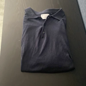 Marin blå långärmad pikétröja från Bläck - En stilren Marinblå långärmad pikétröja från Bläck med klassisk krage och knäppning framtill. Tröjan är tillverkad i ett mjukt material och passar perfekt för en clean och enkel look.