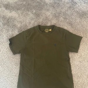 Olivgrön t-shirt från Polo Ralph Lauren - Säljer en olivgrön t-shirt från Polo Ralph Lauren med klassisk blå broderad logga på bröstet. T-shirten har rund halsringning och korta ärmar. Perfekt basic-plagg med snygg passform.
