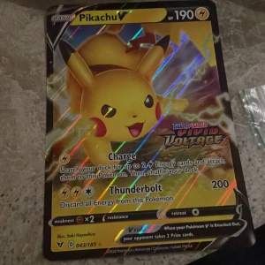 Pikachu V Pokémonkort - Säljer ett glittrande Pikachu V-kort från Sword & Shield Vivid Voltage-serien. Kortet har 190 HP och attacker som Charge och Thunderbolt. Perfekt för samlare eller Pokémon TCG-spelare. Kortnummer 043/185.