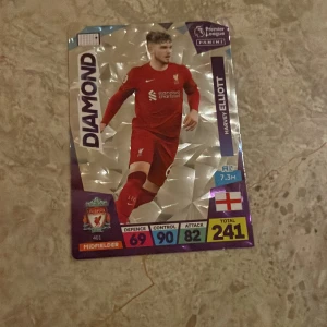 Panini Premier League samlarkort - Harvey Elliott - Säljer ett Panini Premier League samlarkort med Harvey Elliott från Liverpool FC. Kortet har en glittrig bakgrund och visar spelaren i röd matchdräkt. Kortet har statistik och är märkt 'DIAMOND'. Perfekt för samlare!