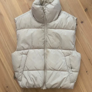 Beige dunväst med hög krage - MÖTS BARA UPP I STOCKHOLM/SÖDERMALM! FRAKTAR INTE   beige dunväst med hög krage och dragkedja framtill. Västen har två snedställda fickor och quiltad design som ger en puffig look. Fläckig.