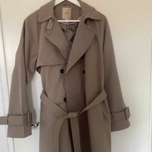 Beige trenchcoat från Vila Rouge - Säljer en klassisk beige trenchcoat från Vila Rouge. Jackan har dubbelknäppning, bälte i midjan och vid ärmsluten samt bred krage. Perfekt för dig som gillar stilrena och tidlösa plagg. Den är helt oanvänd. Nypris 1000kr.