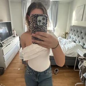 Vit oneshoulder topp med volang - Säljer en vit oneshoulder topp med stor volangdetalj över axeln. Toppen är tight och har en stilren, modern look som passar perfekt till jeans eller kjol.