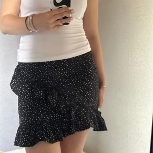 Svart prickig kjol med volang - Supersöt svart kjol med vita prickar och volangdetalj. Kjolen är kort i modellen, perfekt för en trendig look. Lätt och luftig, passar till många olika stilar.😊💓