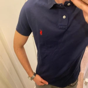 Polo Ralph Lauren (Blå piké) - Snygg blå piké från Polo Ralph Lauren med klassisk krage och knappar framtill. Ikonisk röd broderad logga på bröstet. Perfekt till jeans eller shorts för en stilren look. Den är oanvänd och redo för en ny ägare! Pikén är storlek L!