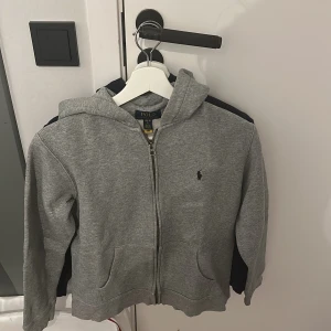 Marinblå hoodie från Polo Ralph Lauren - Säljer en marinblå hoodie från Polo Ralph Lauren med dragkedja och huva. Tröjan har en röd broderad logga på bröstet och två fickor framtill. Perfekt för en avslappnad och stilren look.